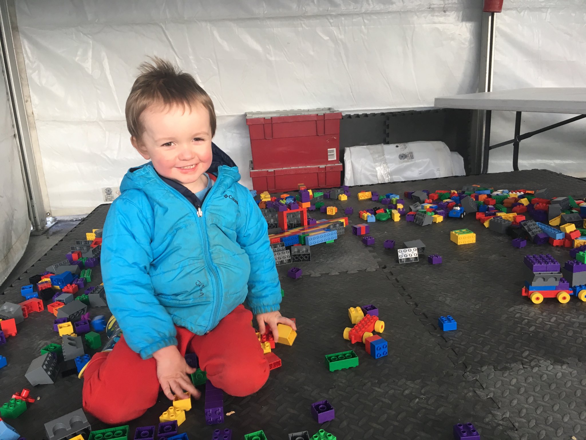 Julian sitting on the ground smiling at the camera dressed in a blue winter coat and red pants. The ground covering is black rubbery tiles and strewn about are brightly colored Duplo bricks.