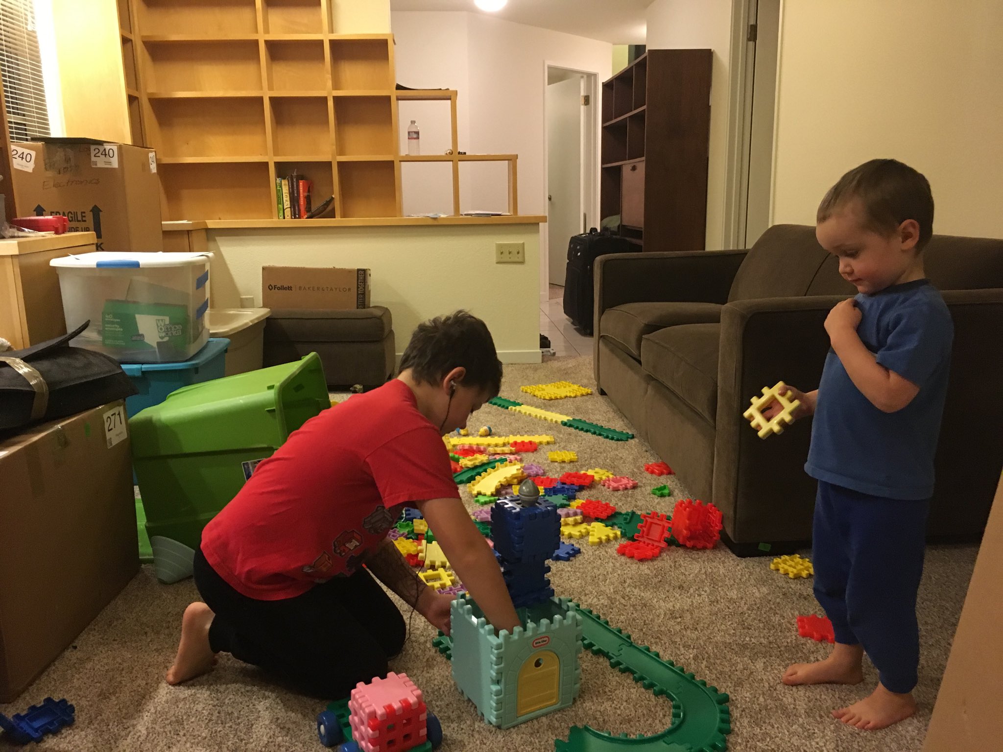 The family room is cluttered with boxes and bins from moving. In the middle of the floor Calvin is playing with waffle blocks while Julian looks on.