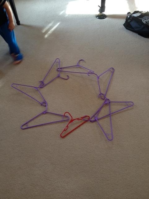 7 purple plastic hangers with one small red plastic hangers hooked together on the beige carpet to form a rough star shape.