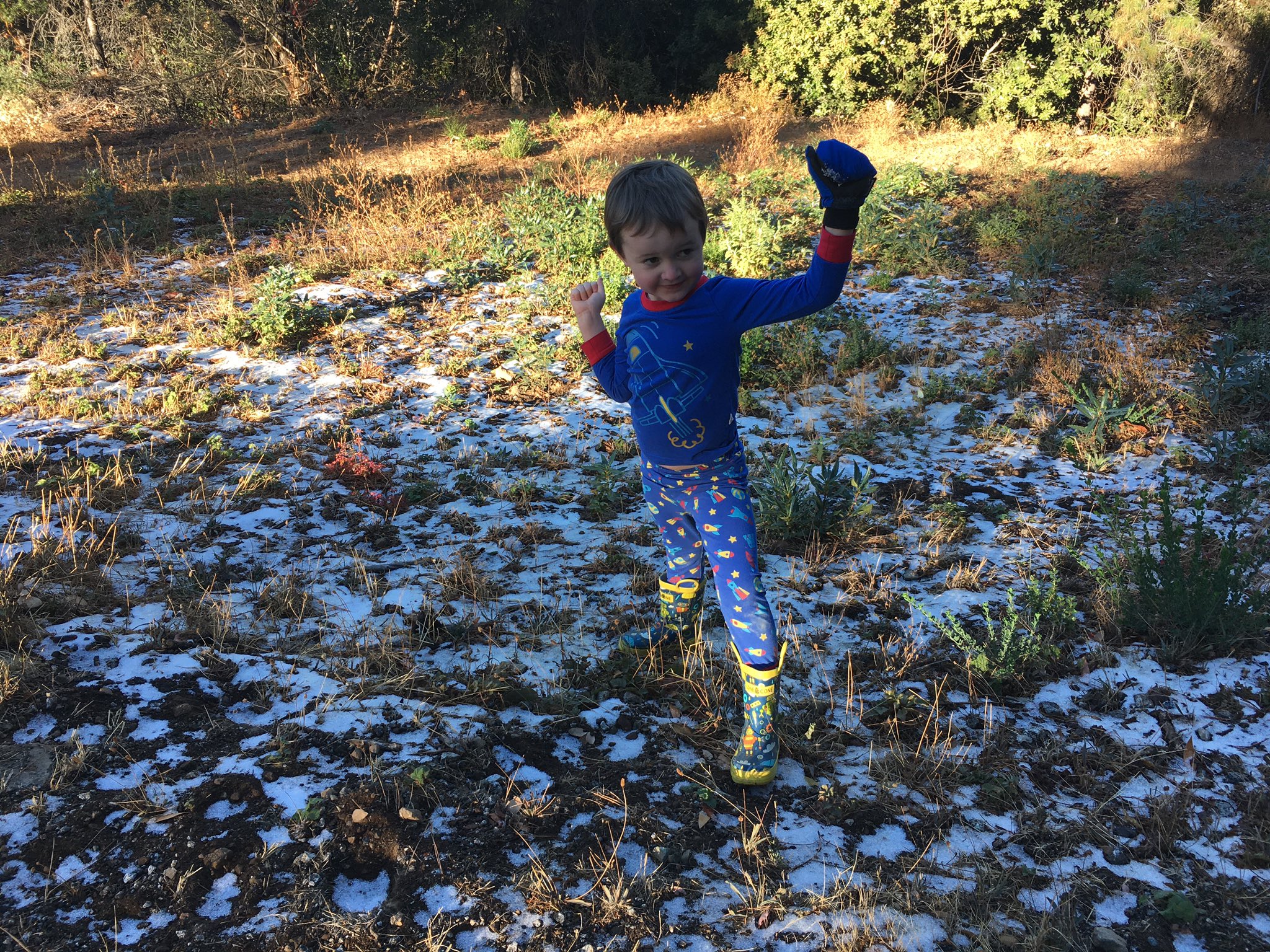 Julian, dressed in pajamas and rain boots, standing on ground lightly spotted with snow.