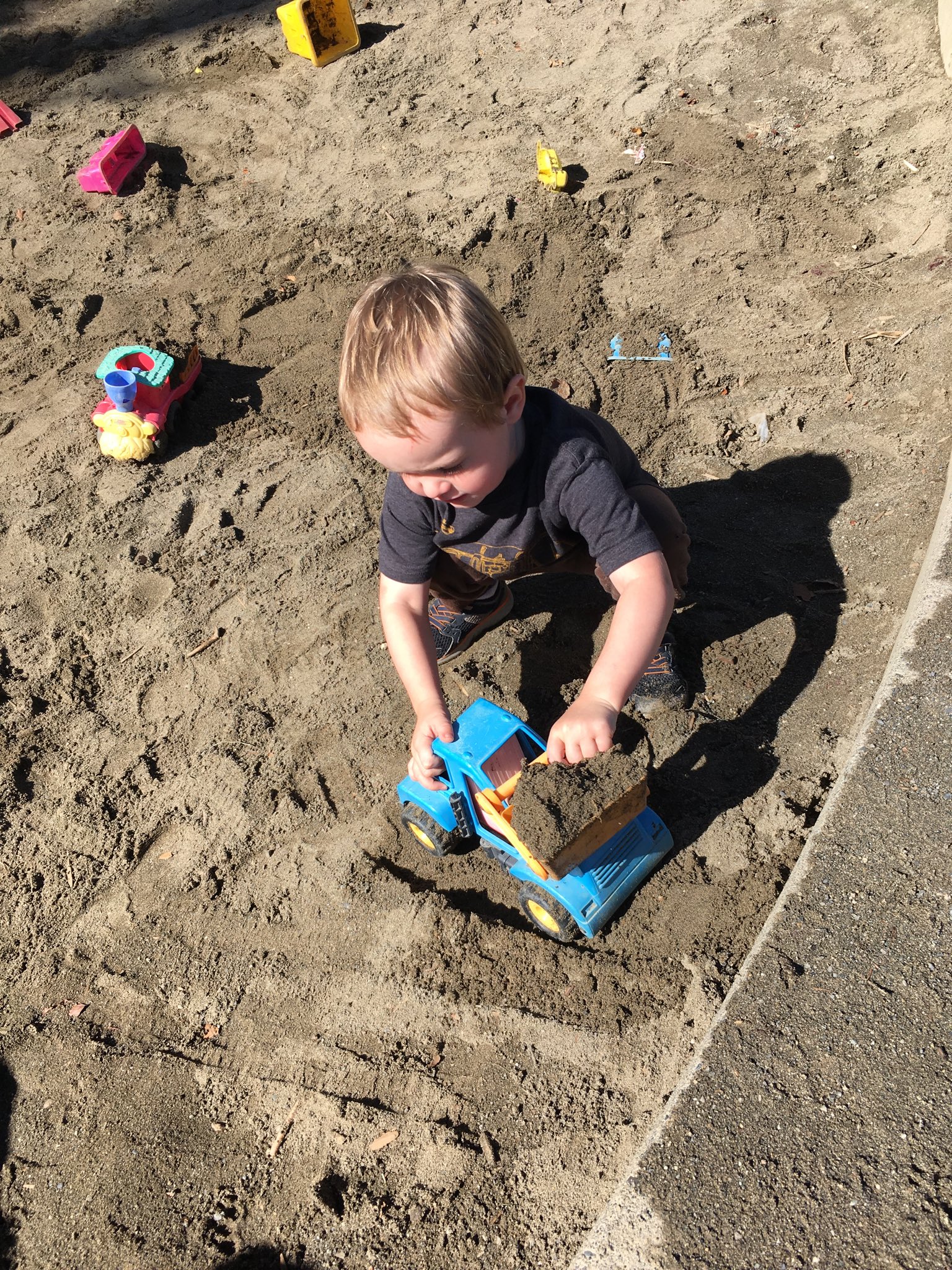 Julian crouches down in a sandbox and plays with a blue plastic construction truck.