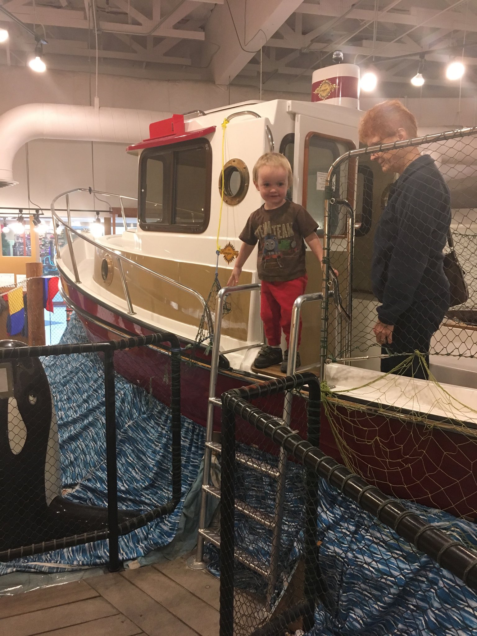 Julian stands on a fishing boat in a museum exhibit. Grandma stands behind him.