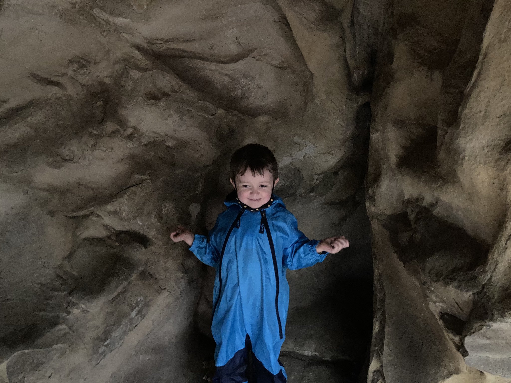 Julian in a cave-like rock formation. The rocks are very pitted. Julian is in a blue rain gear and is holding his hands out on either side, pussibly to catch drips.