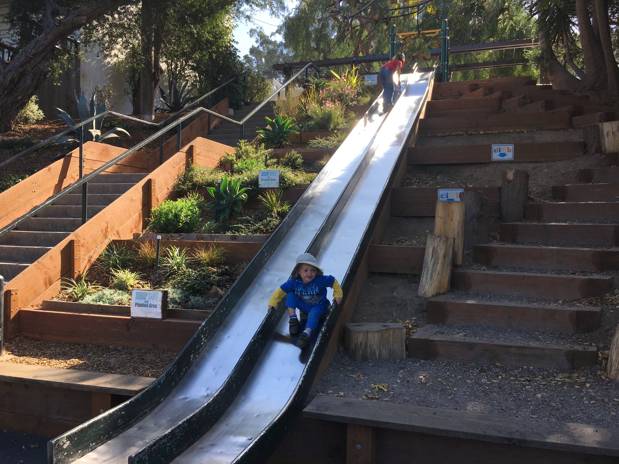 In the middle of the picture are two metal slides next to each other. Julian is almost to the bottom of the slide on the right. To the far left are stairs and in between the stairs and the slide are terraced garden beds. On the right is another set of  stairs made from each and railroad ties.