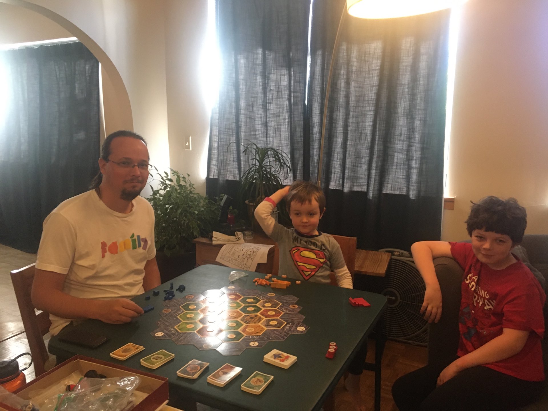 Ted and Julian around a green card table with the hexagonal Catan tiles lade out. Calvin's arm is visible but the rest of him is off screen.