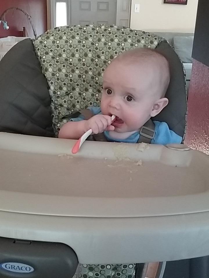 An infant Julian, mostly bald with big eyes, sitting in a high chair putting a spoon into his mouth.