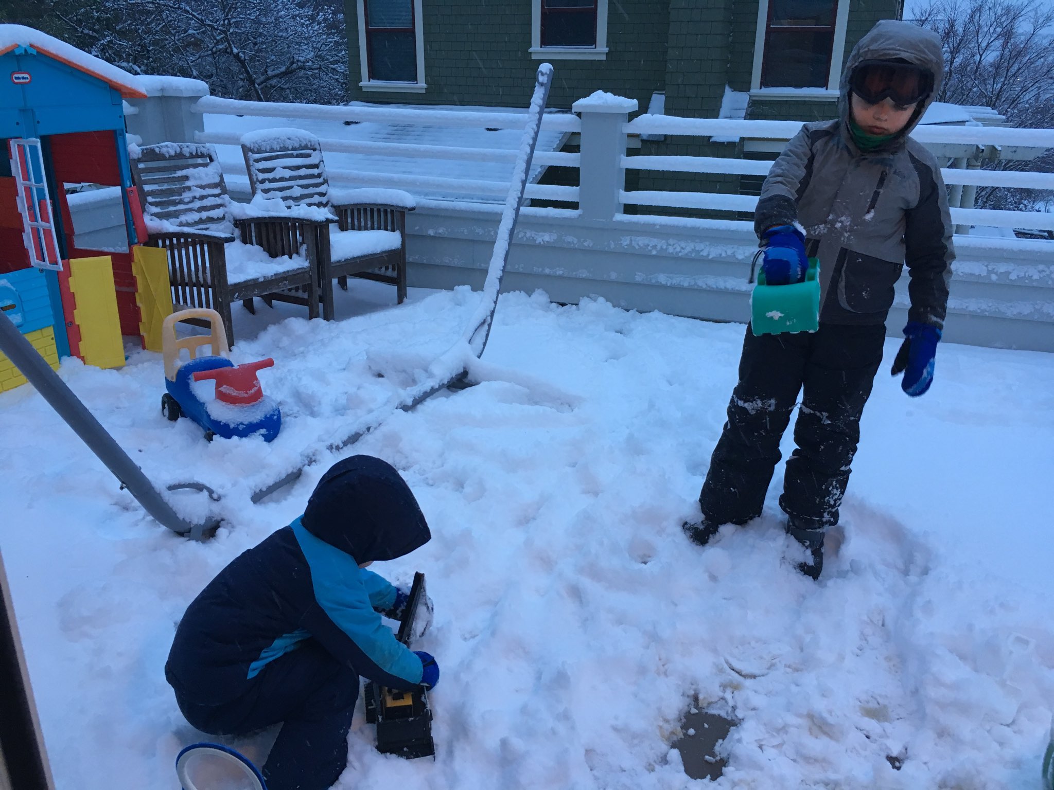 Two kids, bundled up in snow clothes, playing with snow on the deck.