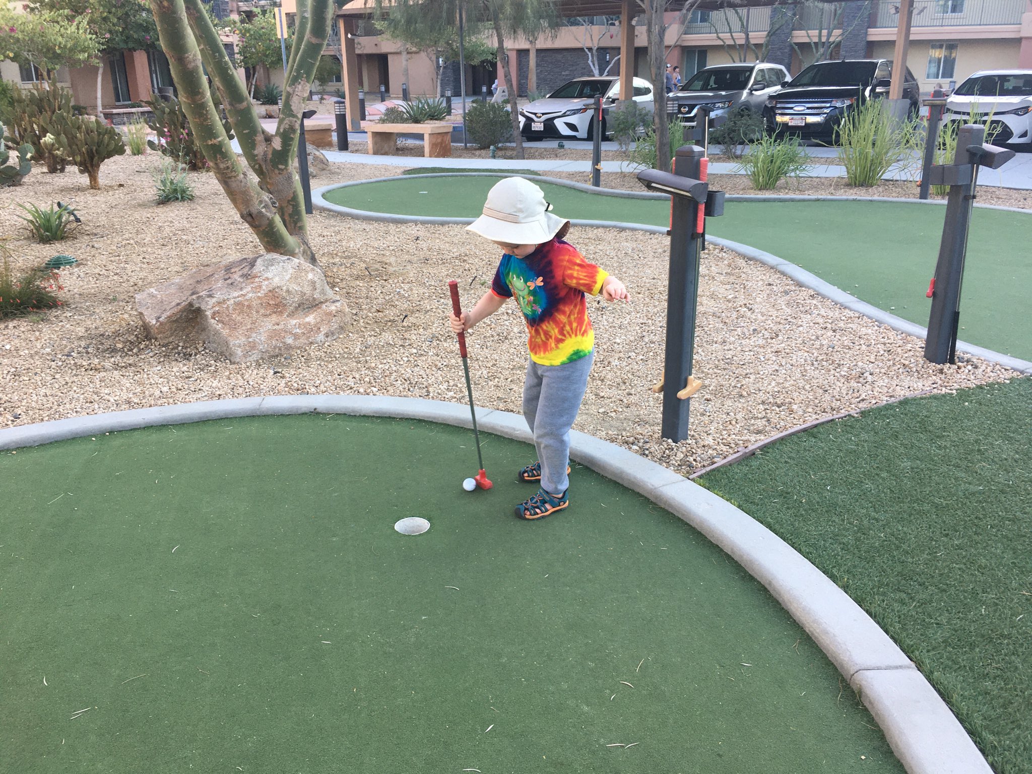 Julian, in a tie-dye shirt and white sun hat, holding a golf putter with his right hand and trying to get a golf ball into a hole.