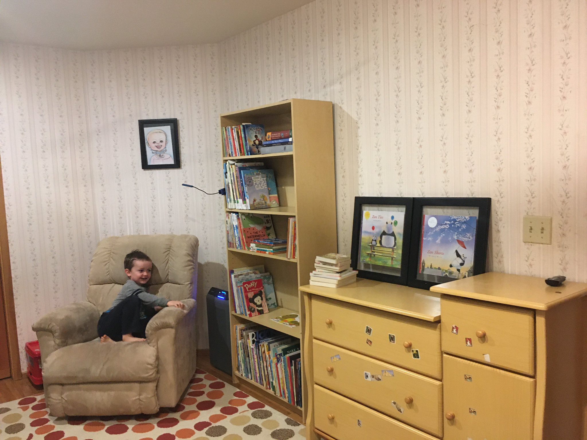 Julian's bedroom. Julian is sitting in a rocking chair. To his right is a bookcase with books and to the right of that is his dresser. Above the rocking chair is a caricature of Julian. The rug on the floor has multi-colored polka dots.