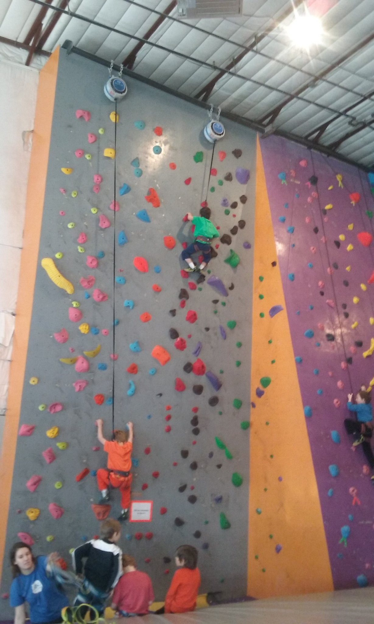 Very tall climbing wall with multi-colored holds. Two kids are on the wall. One is in a orange shirt and then one up further on the right is in a green shirt.