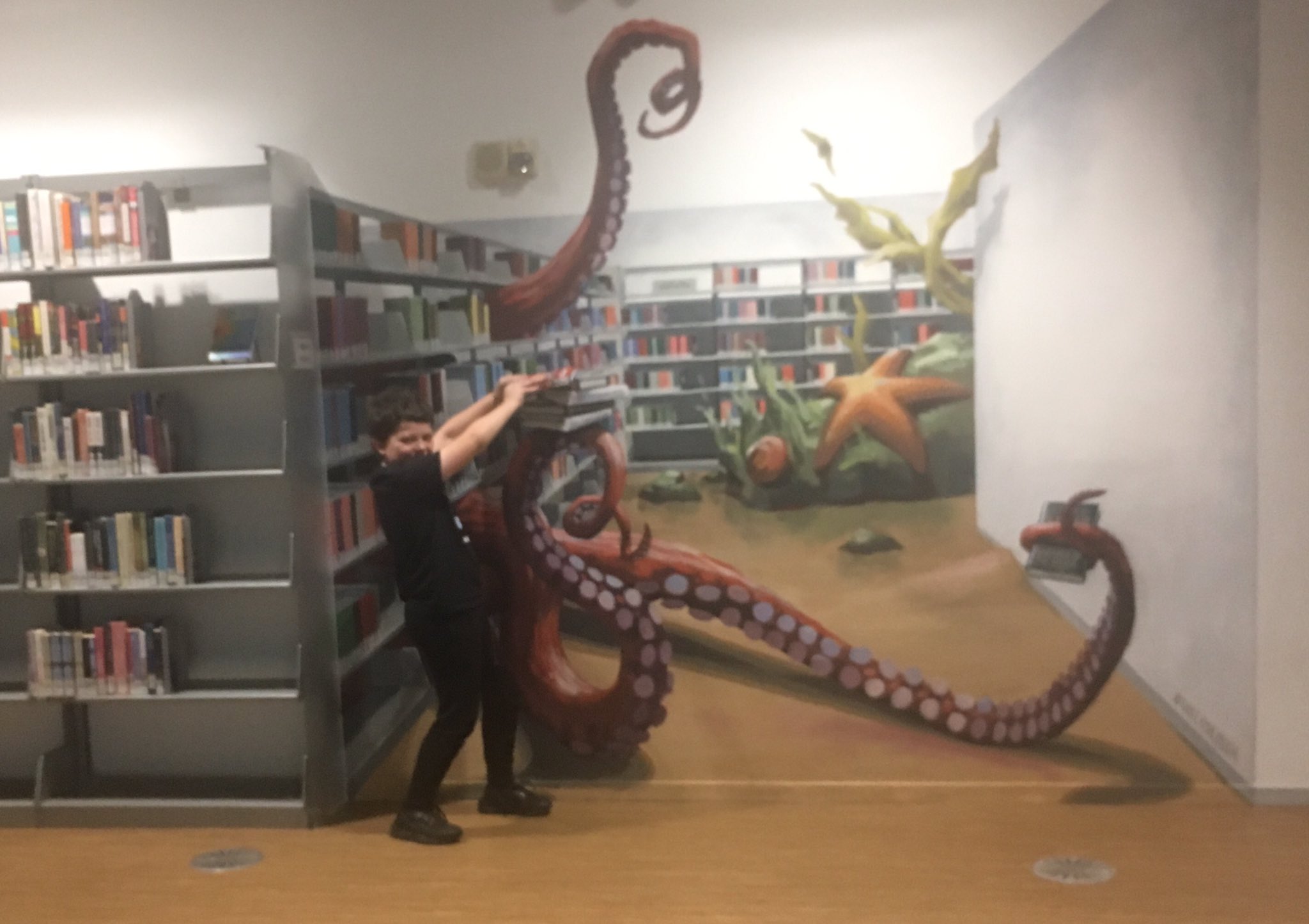 Off to the left is a library bookshelf. To the right of the bookshelf is a mural painted to look like additional library books stacks and it looks like very large octopus arms are reaching through the open shelving. Calvin is pretending to try pulling a book away from the octopus.