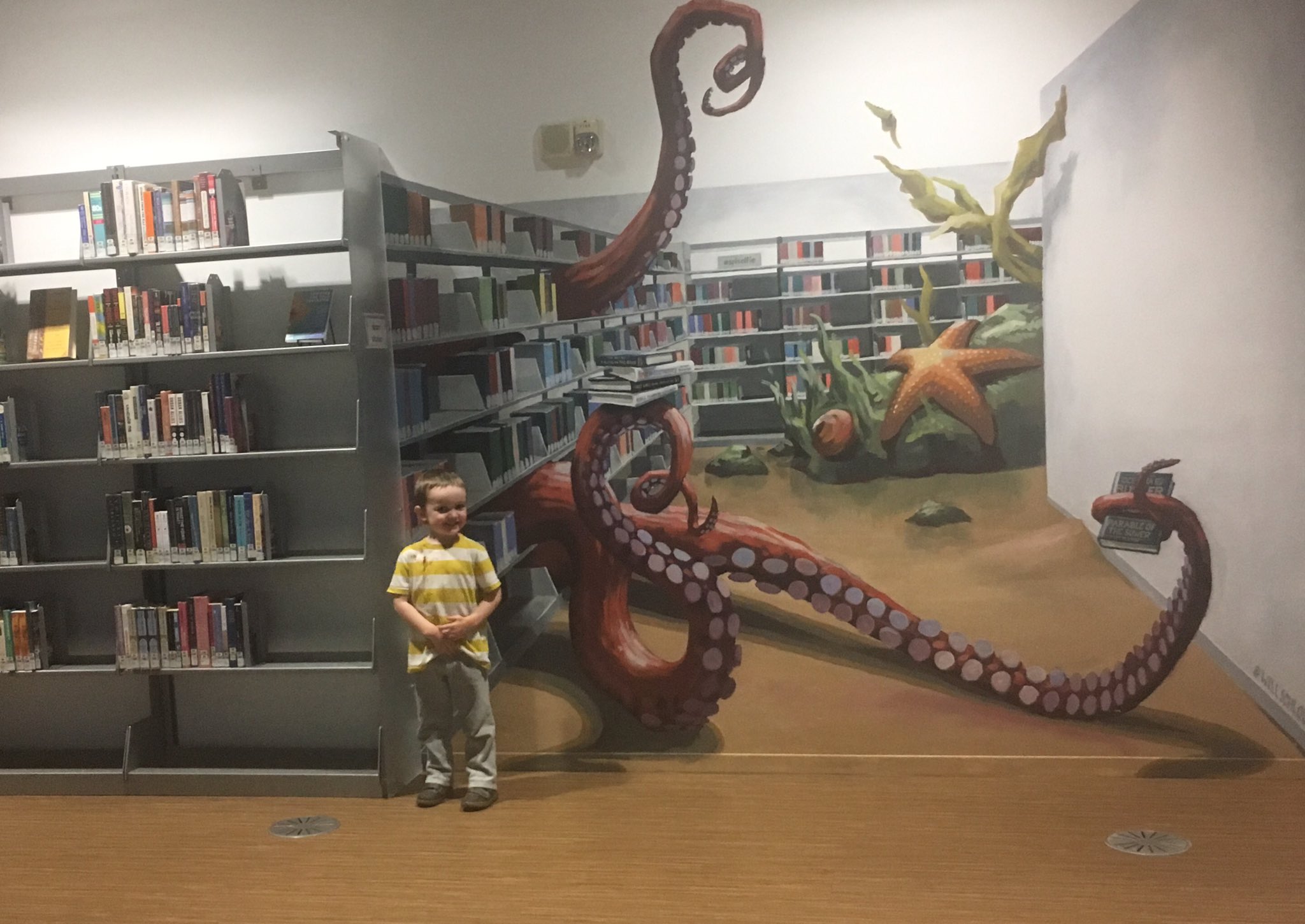 Off to the left is a library bookshelf. To the right of the bookshelf is a mural painted to look like additional library books stacks and it looks like very large octopus arms are reaching through the open shelving. Julian is standing next to the octopus arms.