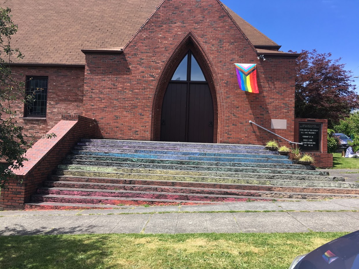 Church with rainbow colored steps, one color per step, and flying a progressive pride flag.