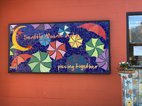 Mosaic on red wall. Blue background with a yellow crescent moon and multi-colored umbrellas. Says Seattle Mosaic Arts piecing together.