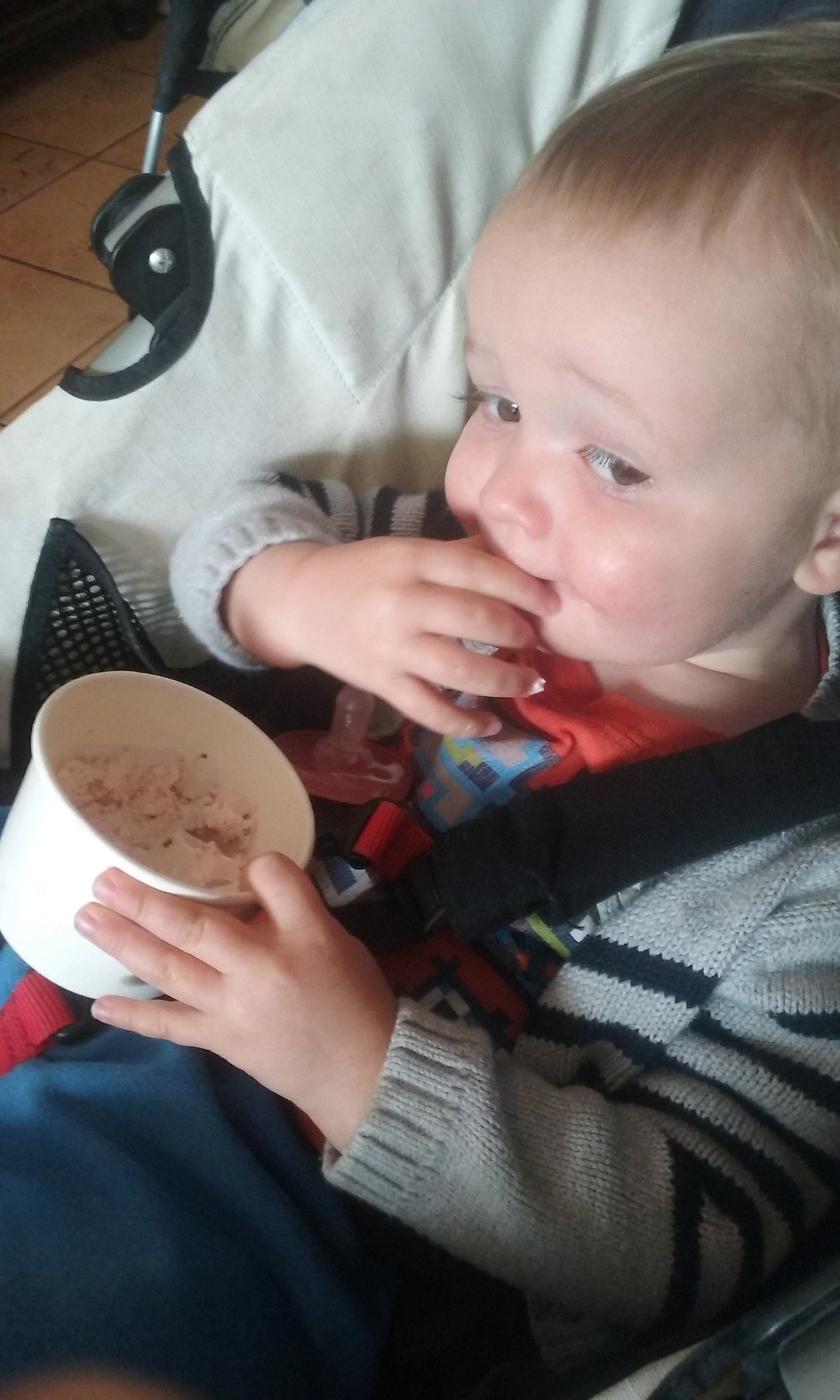 Julian, strapped into a stroller, holds a paper bowl of ice cream in his left hand while sucking the fingers on his right hand.